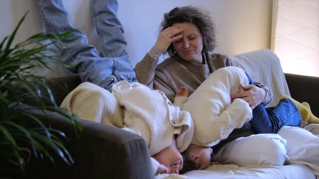 Exhausted mother experiencing a headache while her two hyperactive twin children are uncontrollably jumping upside down on the sofa in the living room, a challenging moment in motherhood