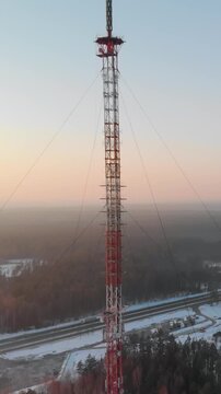 Vertical drone shot of a tall red and white steel lattice broadcasting tower. Communication infrastructure standing above winter forest and highway during golden hour