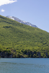 Fototapeta premium vibrant green mountain slopes meet the deep blue waters of a serene alpine lake under a clear sky, featuring distant snow-capped peaks and lush spring vegetation
