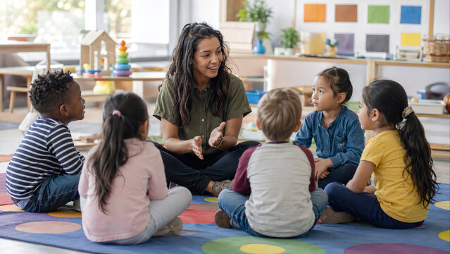 Cheerful female teacher sitting in a circle with a diverse group of young children on a colorful rug in a bright preschool classroom, engaging in a storytelling session and lesson.