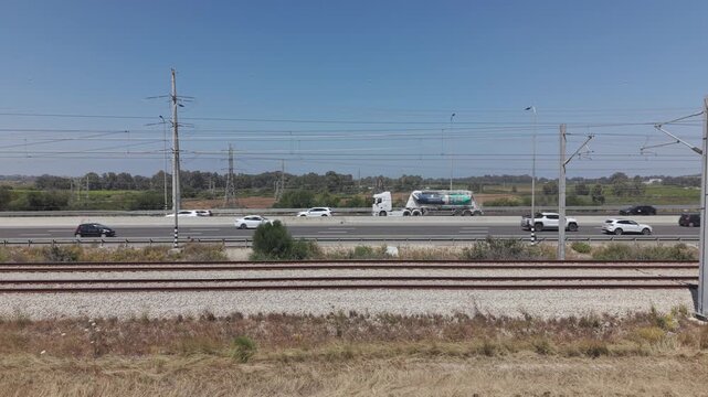Double railway tracks in foreground alongside busy highway with moving cars and catenary poles in Israel
