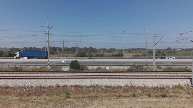 Railway tracks in foreground with busy multi-lane highway traffic and power lines across flat landscape in Israel
