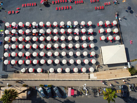 Aerial view of rows of white umbrellas and red sunbeds on a dark pebble beach with people walking and parked cars along the promenade in Imperia, Liguria, Italy.