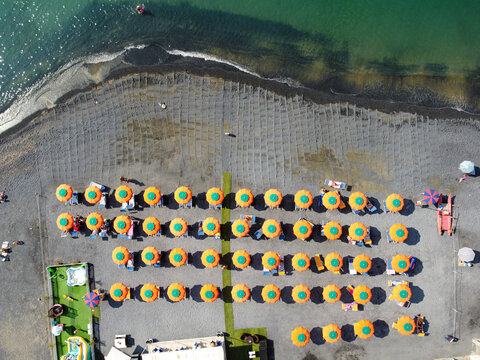 Aerial view of rows of orange and green sun umbrellas on a grey pebble beach by the turquoise sea in Imperia, Liguria, Italy.