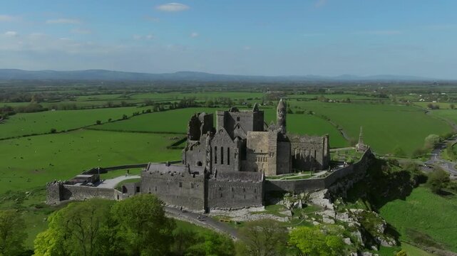 Smooth drone fly around of Rock of Cashel with panoramic countryside views and vibrant green farmland beneath bright summer skies.