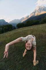 Fototapeta premium Young woman performs a backbend in a grassy meadow with distant mountains, capturing a playful, adventurous moment outdoors.