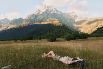 Fototapeta premium Young woman lies in a sunlit meadow with mountains in the background, relaxed pose and peaceful expression amid tall grass, a gentle breeze, warm light, natural colors, distant pines, and wide open