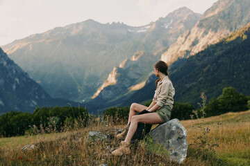 Fototapeta premium Young woman sits on a rock in a meadow with a distant mountain panorama, enjoying peaceful solitude in nature and feeling calm and free.