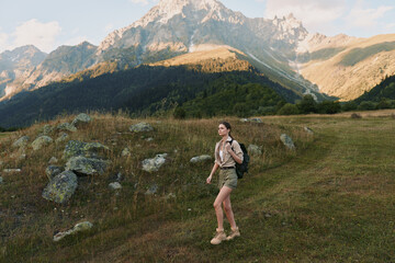 Fototapeta premium Woman hiker walking through a meadow beside rocky mountains, carrying a backpack, enjoying outdoor nature and expansive scenery in daylight