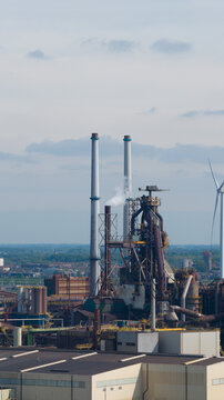 Aerial view of Tata Steel industrial plant featuring blast furnaces, tall chimneys emitting steam, and a wind turbine under a cloudy sky in Velsen-Noord, Noord-Holland, Netherlands.