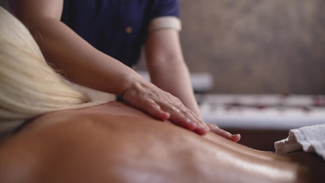 Close up view shows masseuse giving relaxing back massage to blonde woman. Oiled skin glistens under soft light. Shallow depth of field creates serene, therapeutic mood