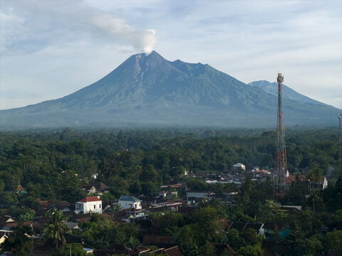 Aerial view of Mount Merapi volcano with smoke rising from the crater overlooking a village with a telecommunications tower and lush green trees in Klaten, Central Java, Indonesia.