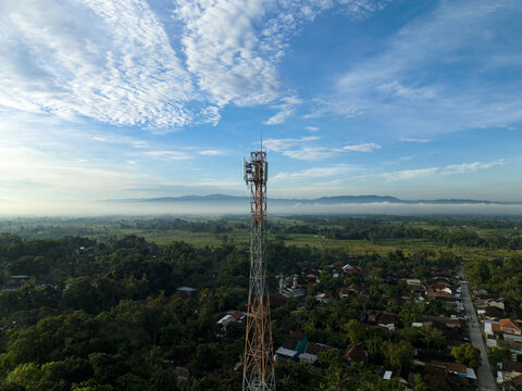 Aerial view of a tall telecommunications tower rising above a rural village surrounded by lush green trees and misty mountains under a blue sky in Klaten, Central Java, Indonesia.