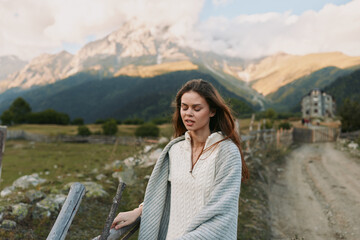 Fototapeta premium A young woman with brown hair walks along a rustic fence beside a mountain tapestry wrapped in a light knit sweater calm and thoughtful in the open countryside