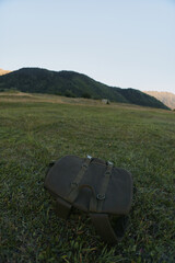 Obraz premium A single rustic satchel rests on a green grass field in a sunlit meadow, suggesting an outdoor escape and quiet travel mood during a calm day.