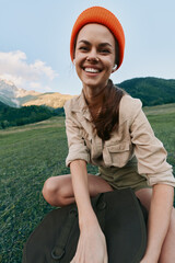 Fototapeta premium Cheerful woman lifestyle outdoors, smiling on a sunlit mountainside field, casual beige shirt and orange beanie, capturing a carefree moment with nature and wide landscape