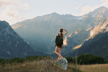 Fototapeta premium Young woman with backpack stands on a rock in a mountain valley, looking toward distant peaks in a sunlit alpine landscape, feeling freedom and adventure as a gentle breeze rustles grass