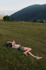 Fototapeta premium Relaxed woman lying on grass in a wide countryside field with distant mountains, capturing a casual outdoor lifestyle moment of rest, freedom, and connection with nature.