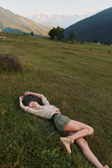 Fototapeta premium Young woman lies on a grassy hillside in a mountain valley, relaxing and enjoying nature with warm sunlight, casual outfit and a peaceful mood, soft breeze rustling grass as distant peaks glow.
