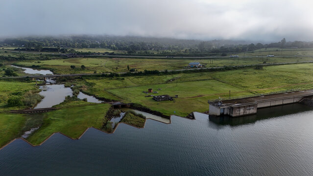 Aerial view of Midmar Dam wall with green fields, a winding river, and misty hills under an overcast sky in Howick, KwaZulu-Natal, South Africa.