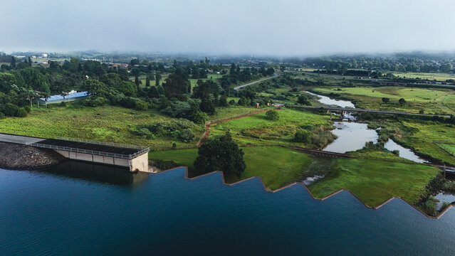 Aerial view of Midmar Dam featuring its unique zigzag spillway wall, surrounded by lush green fields and misty hills in Howick, KwaZulu-Natal, South Africa.