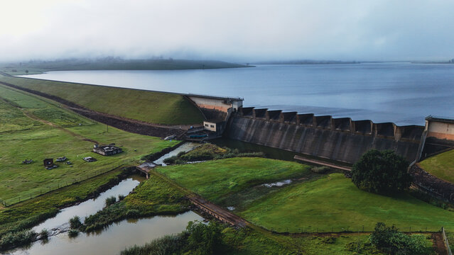Aerial view of Midmar Dam featuring the concrete spillway and reservoir surrounded by green hills under a misty sky in Howick, KwaZulu-Natal, South Africa.