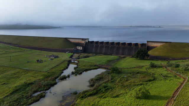 Aerial view of Midmar Dam featuring its concrete spillway and grassy embankments under misty hills and a cloudy sky in Howick, KwaZulu-Natal, South Africa.