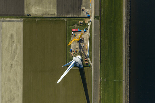 Aerial view of a large wind turbine under maintenance with a yellow mobile crane and support vehicles in a flat agricultural landscape next to a canal with a tractor in the distance.