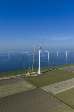 Aerial view of wind turbines being maintained by a large yellow crane on a coastal field next to the sea under a clear blue sky.