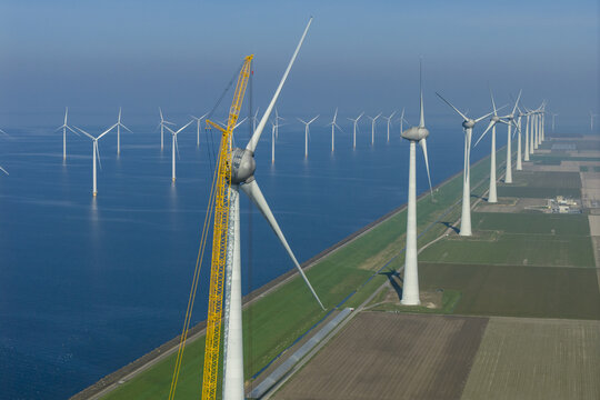 Aerial view of a row of offshore and onshore wind turbines with a tall yellow maintenance crane along a coastal dike and flat agricultural fields under a clear blue sky.