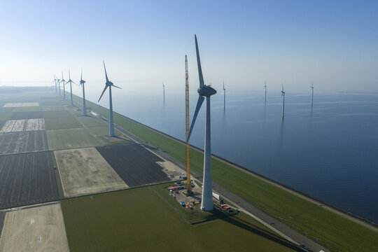 Aerial view of a row of modern wind turbines standing along a coastal dike and in the water between agricultural fields and the sea.