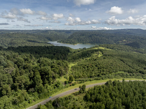 Aerial view of Chemosusu Dam surrounded by dense green forests and rolling hills with cars driving on a road in Chemosusu, Baringo County, Kenya.
