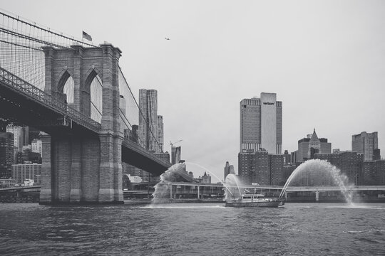 New York, USA, 2 May 2026: Monochrome FDNY fireboat water salute display on East River NYC. Black and white wide shot of fireboat spraying water jets near Brooklyn Bridge and Lower Manhattan.