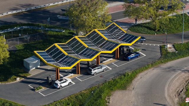 Bant, Netherlands - 23 April 2026: Aerial view of Fastned Charging Station featuring a yellow solar panel roof with electric cars parked in charging bays surrounded by green trees.
