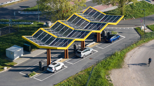 Bant, Netherlands - 23 April 2026: Aerial view of Fastned charging station with its iconic yellow solar roof, electric cars parked at charging points, and a cyclist on the road.