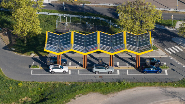 Bant, Netherlands - 23 April 2026: Aerial view of Fastned Charging Station with its iconic yellow zig-zag solar roof and electric vehicles parked in charging bays surrounded by green trees.