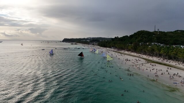 Malay, Philippines - 02 May 2026: Aerial view of Boracay Island featuring sailboats on turquoise water and people on the white sand beach lined with palm trees under a cloudy sky.