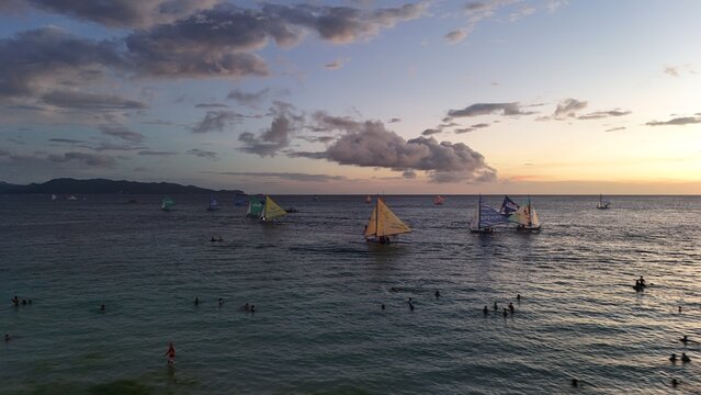 Malay, Philippines - 02 May 2026: Aerial view of Boracay Island with traditional paraw sailboats on the calm sea at sunset, featuring people swimming and a cloudy sky.