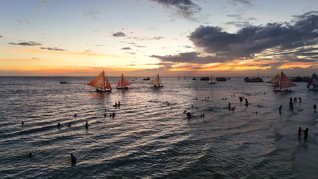 Malay, Philippines - 02 May 2026: Aerial view of Boracay Island with people swimming in the sea and traditional paraw sailboats under a vibrant sunset sky.