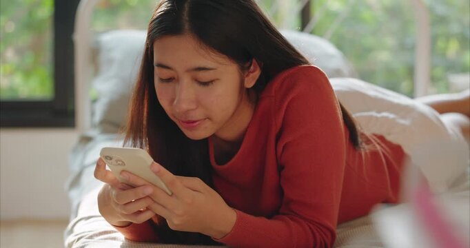 young Asian woman lies on a neatly made bed, smiling as she uses a light case smartphone. Soft daylight illuminates a cozy bedroom. 