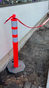 A flexible delineator round post with base placed next to a low white wall, with orange caution tape, marking area of rough, incomplete, pavement work. In vertical format.