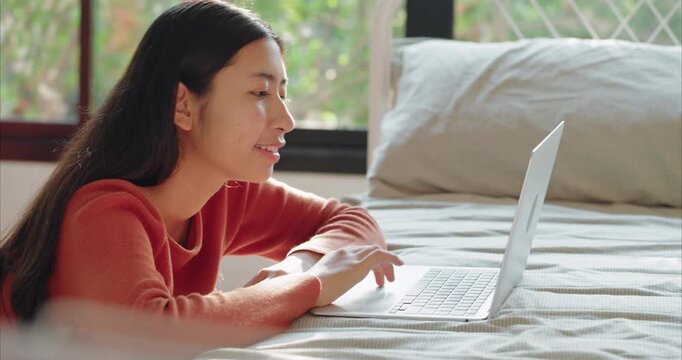 Asian Teenage Girl in Red Shirt Using Laptop on Bed, 
 in Cozy Bedroom with Warm Morning Sunlight, Side View.