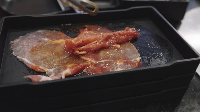 Raw marinated beef slices on a black plastic tray being handled with metal tongs in a restaurant. Perfect for Korean BBQ, sukiyaki, or grilled meat buffet concepts. Fresh ingredients ready.