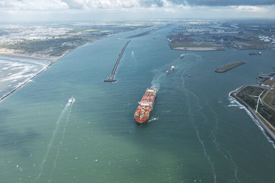 Rotterdam, Netherlands - 05 September 2019: Aerial view of a large container ship entering the port through the breakwaters with industrial facilities and a wind turbine under a cloudy sky.