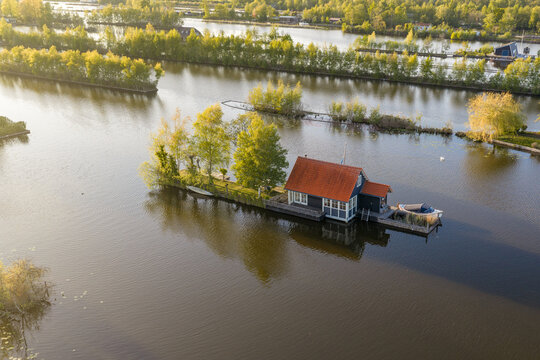 Breukelen, Netherlands - 25 April 2026: Aerial view of Scheendijk recreation area featuring a small island with a wooden house and lush trees surrounded by calm water during golden hour.