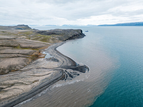 Svalbard and Jan Mayen - 17 July 2025: Aerial view of the rugged coastline with sediment plumes flowing into the turquoise arctic waters and a ship in the distance.