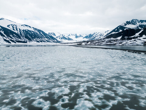 Svalbard and Jan Mayen - 02 July 2025: Aerial view of a frozen arctic fjord with intricate ice patterns on the water surface and snow-capped mountains under an overcast sky.