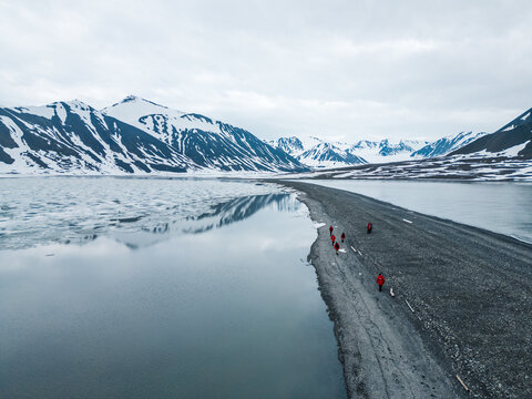 Svalbard and Jan Mayen - 02 July 2025: Aerial view of people in red jackets walking along a narrow gravel spit between icy waters and snow-capped mountains.