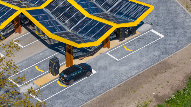 Bant, Netherlands - 21 April 2026: Aerial view of Fastned Charging Station with its iconic yellow honeycomb solar roof and an electric car parked at a charging point on a sunny day.