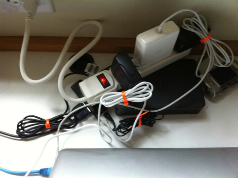 A collection of tangled electrical cables, chargers, and a power strip on a desk, with a laptop and wall socket visible in the background